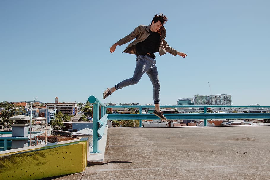 man jump from blue fence to brown concrete pavement, photography of man wearing brown jacket in open air HD wallpaper
