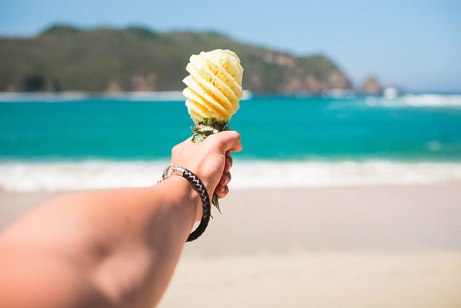selective focus photography of person holding fruit near beach, shallow photo of person holding yellow pineapplae HD wallpaper