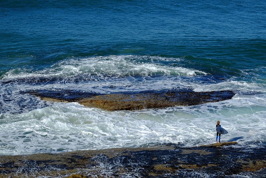 person about to surf on shore, person standing on rock surrounded of ocean during daytime HD wallpaper