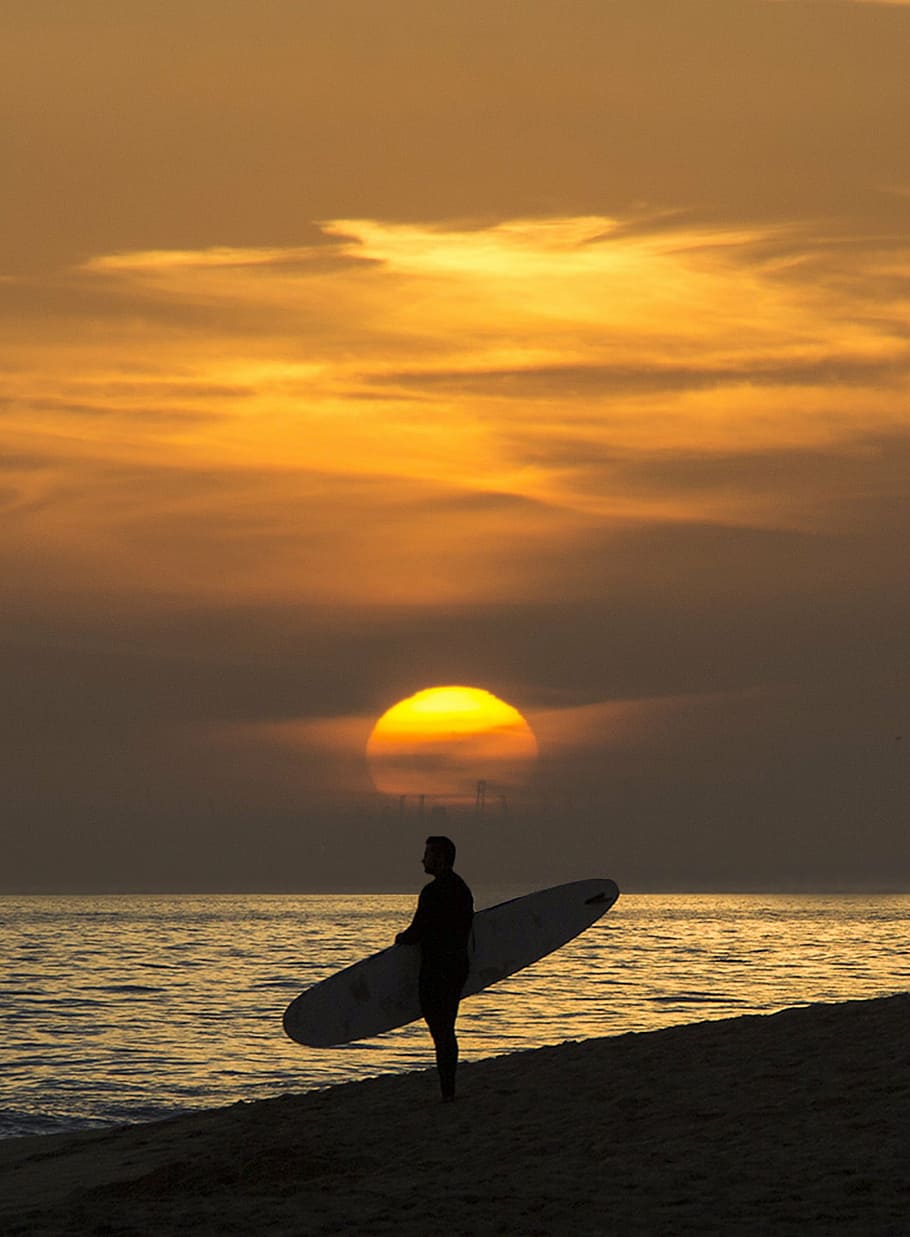 silhouette photo of man standing on seashore while holding surfboard during golden hour, HD wallpaper