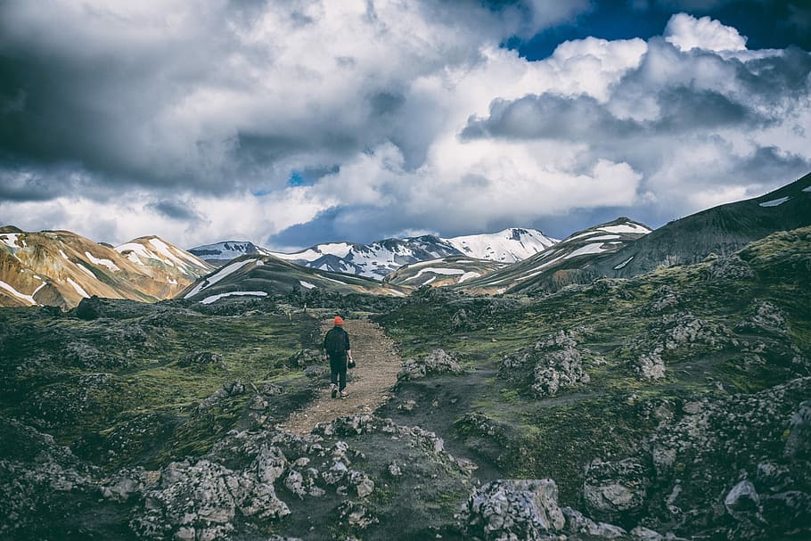 person in black suit walking on brown road, person wearing black top walking on brown road surrounded with gray rocks and green grass under white clouds and blue sky during daytime HD wallpaper