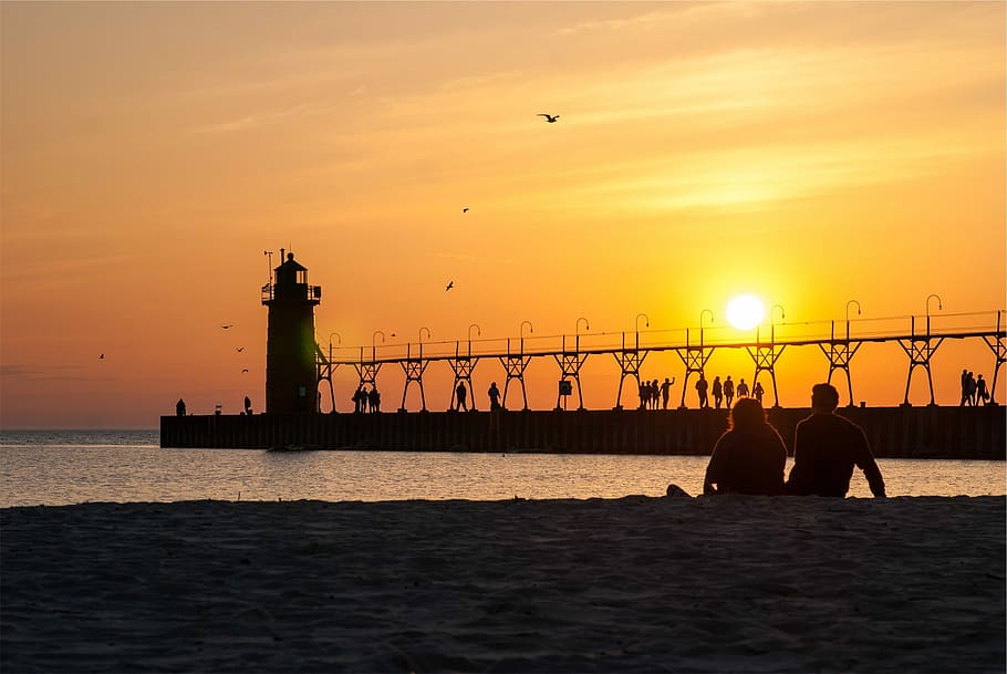 silhouette photo of man and woman sitting on shoreline during golden hour HD wallpaper
