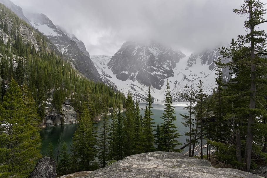 gray rock mountain leading to river and coated snow mountain, clouds covering peak of mountain near body of water and trees HD wallpaper
