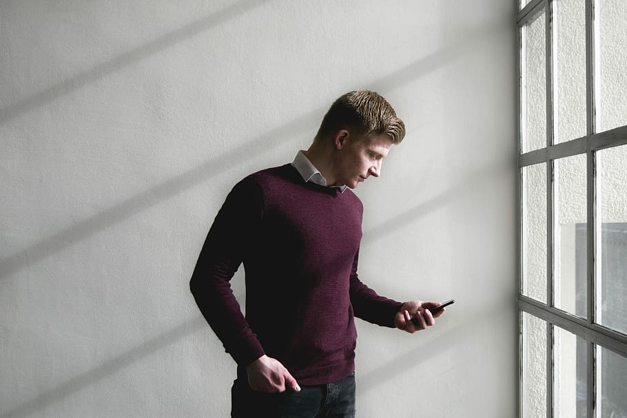 man standing near white concrete wall while using smartphone, man holding smartphone near window HD wallpaper