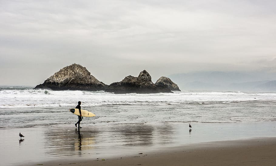 Man Carrying Surfboard in Front Bodies of Water on Brown Sand HD wallpaper