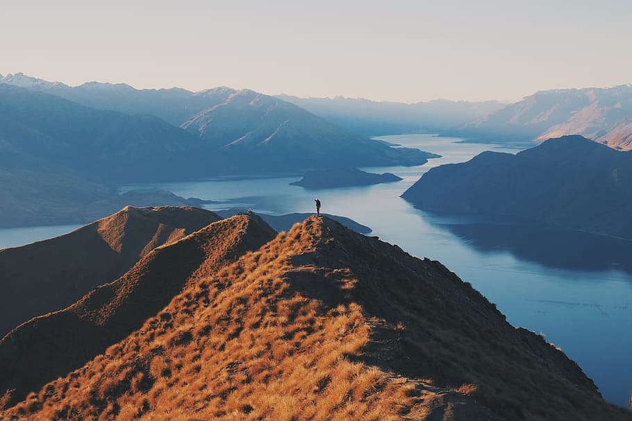 person standing on top of hill, brown rocky mountain near water photo during daytime HD wallpaper