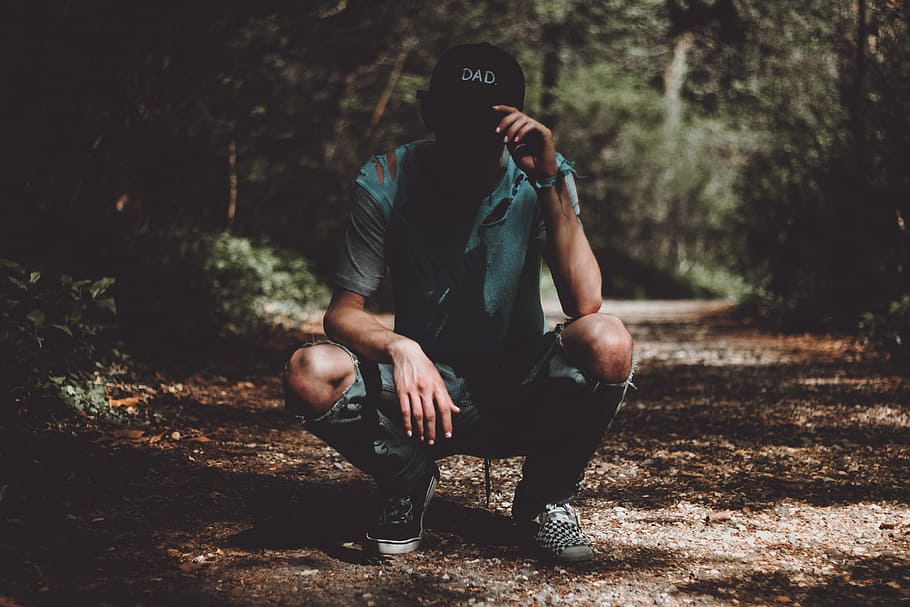 man holding cap at the pathway near trees, man sitting on brown soil during daytime HD wallpaper