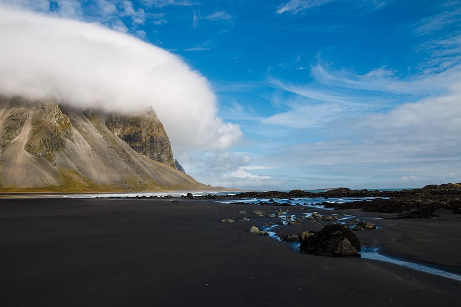 mountain hill covered with clouds near body of water during daytime, black sand near body of water during daytime HD wallpaper