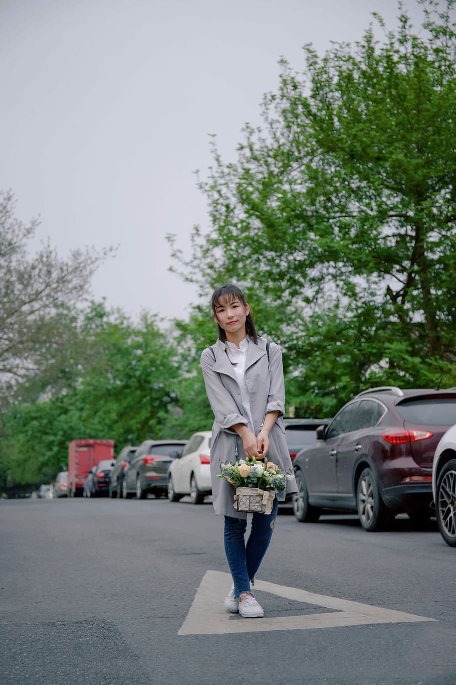 woman holding basket of flower standing in middle of road near parked vehicles, woman holding basket standing near vehicles on road under tree at daytime