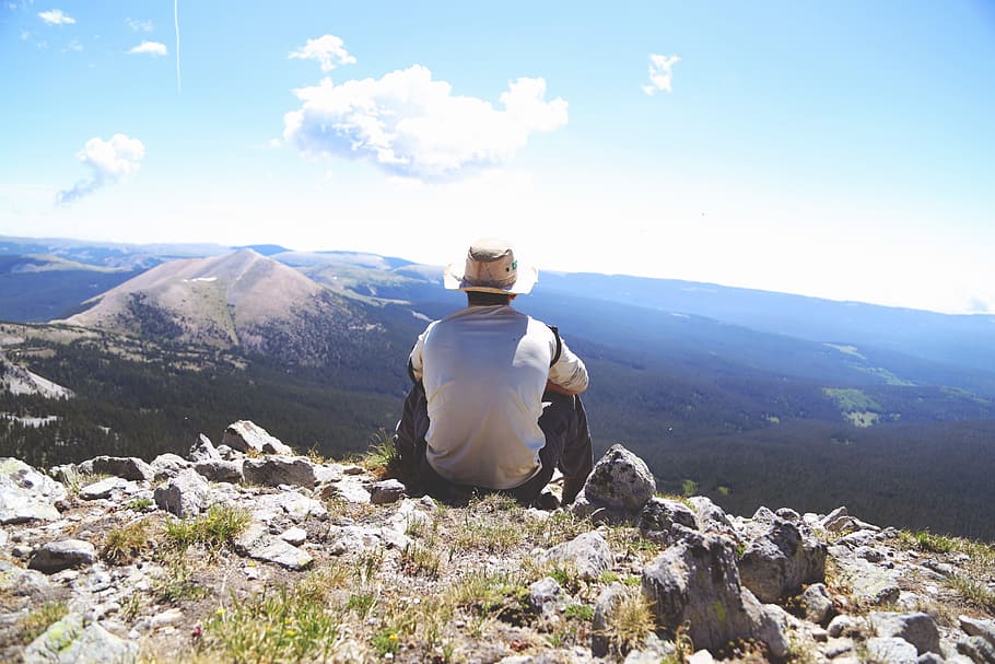 person sitting on a gray rock watching over a mountain, man in white top sitting on rocks
