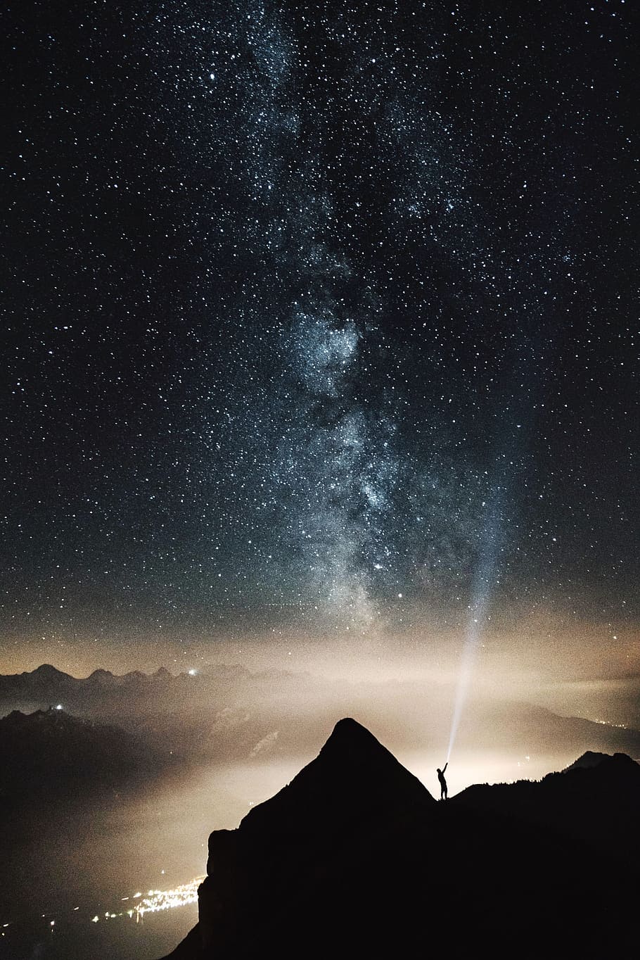 silhouette of person on top of mountain pointing flashlight on sky filled with stars at night time, person holding flashlight on mountain
