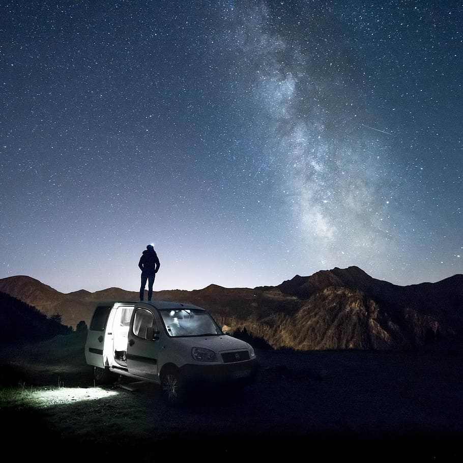 man standing on white minivan looking at mountain under milkyway, silhouette of person standing on white van