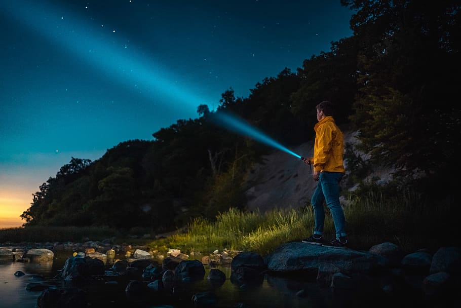 man holding flashlight standing on gray stone during nighttime, man in yellow jacket using flashlight in front of body of water HD wallpaper