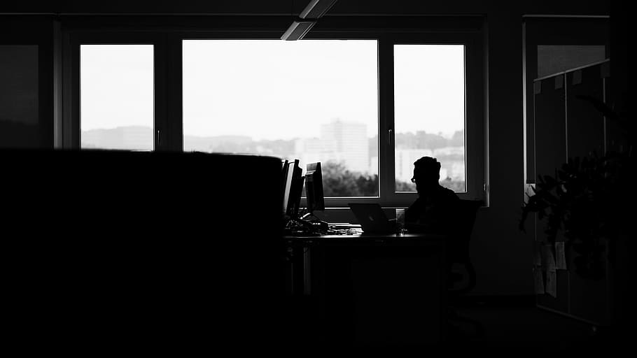 silhouette of man sitting along glass window, photo of man on black desk using laptop