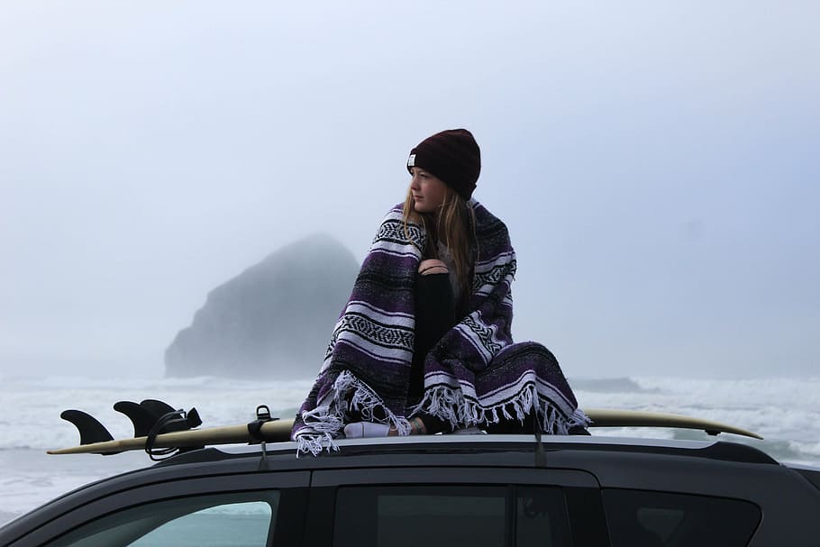 woman with blanket sitting on surfboard on top of vehicle near sea in distant of mountain isle HD wallpaper