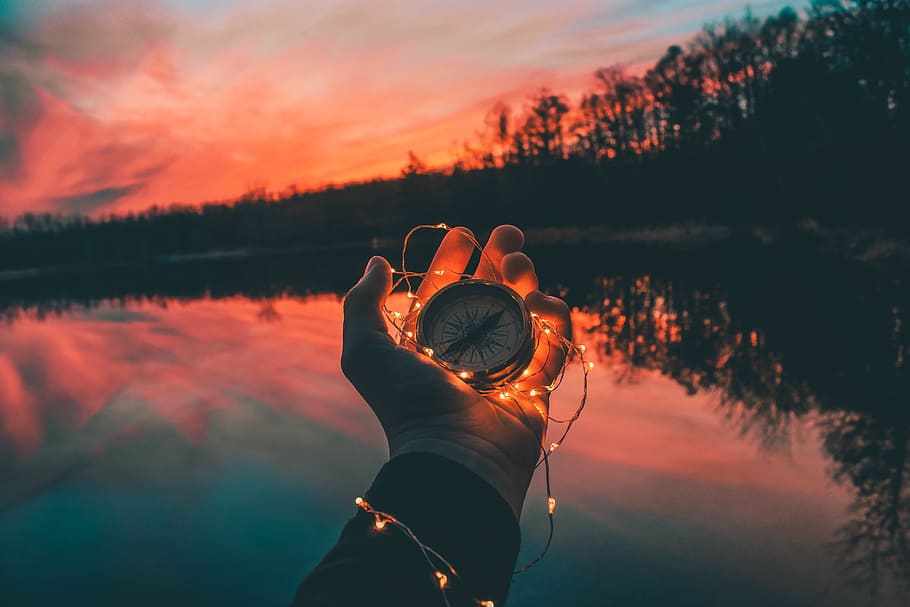 person holding compass and string light near body of water during sunset HD wallpaper