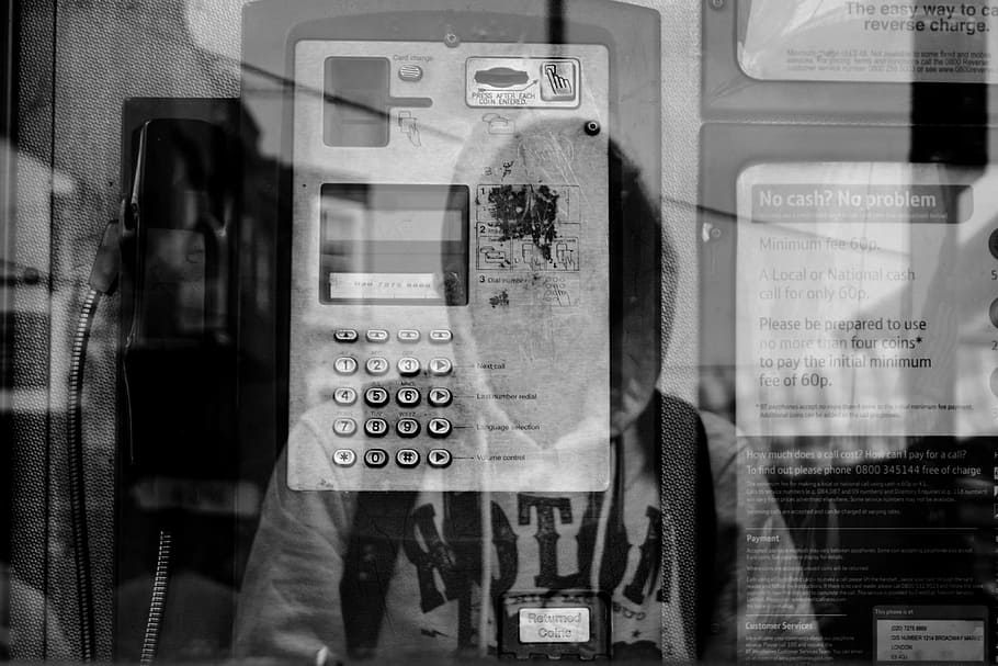 grayscale photo of reflection of person wearing hoodie carrying backpack on glass telephone booth wall, grayscale photo of telephone booth