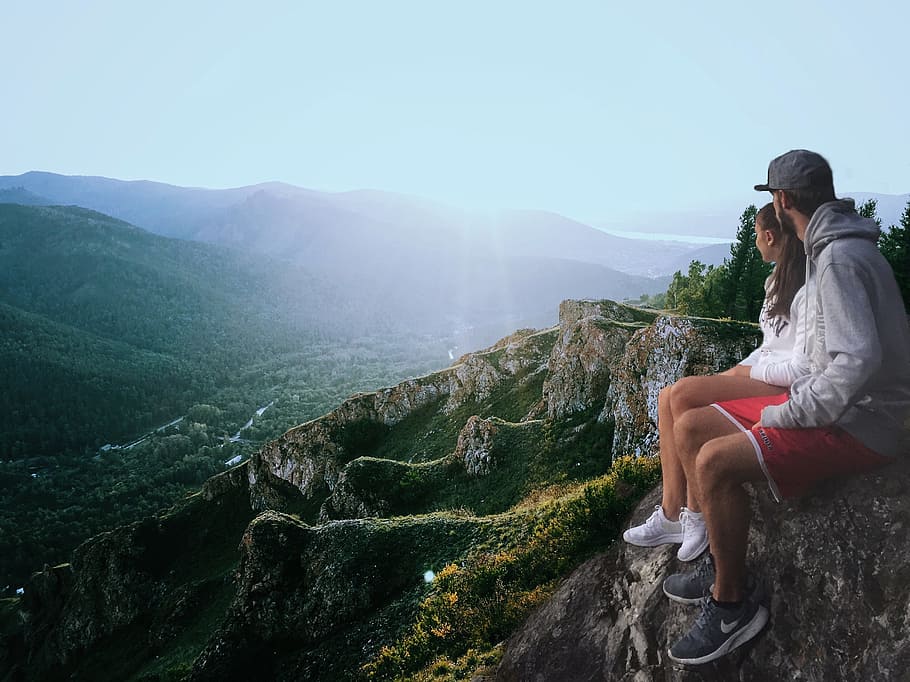 couple sitting on edge while looking at the mountains, man and woman sitting on cliff overlooking sun rise over the hills