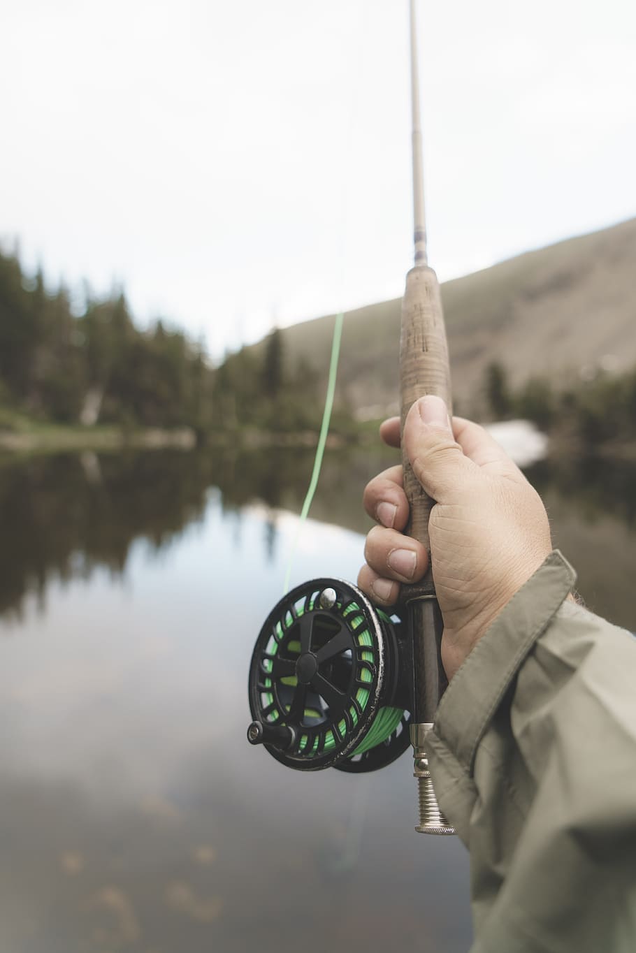 shallow focus photography of person holding black and brown fishing rod, man holding brown and green fishing rod standing near lake under white sky