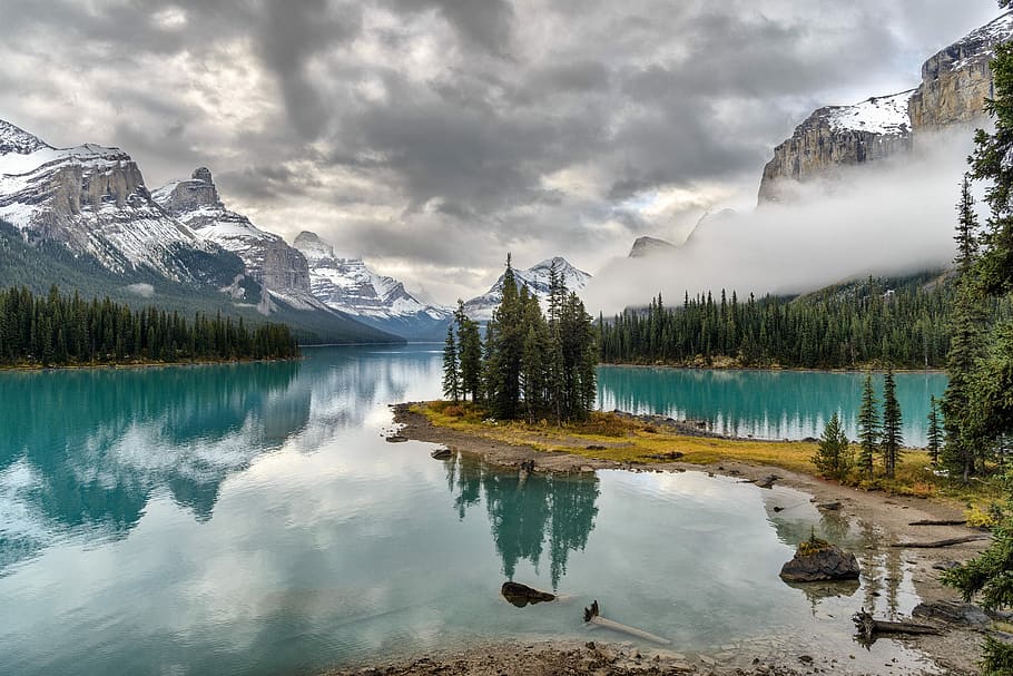 body of water surrounded by pine trees at the foot of snow covered mountain during daytime, lake surrounded by mountain ranges HD wallpaper