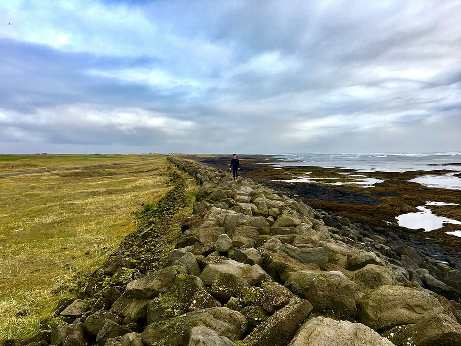 person walking on rocks near body of water under cloudy sky, person standing on sea shore HD wallpaper