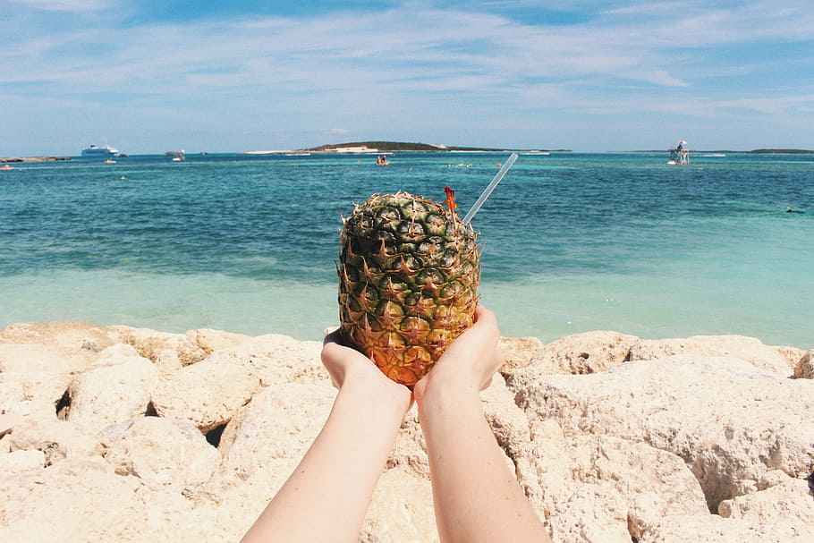person holding pineapple on seashore during daytime, person holding pineapple with straw on beach shore HD wallpaper