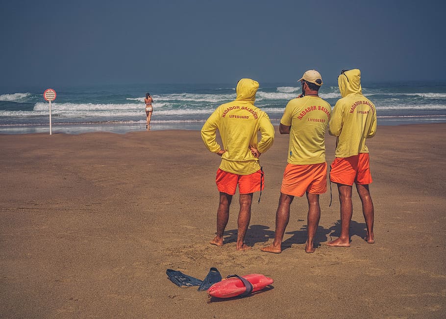 three men standing on brown sand starring in woman standing at seashore, three lifeguard at beach