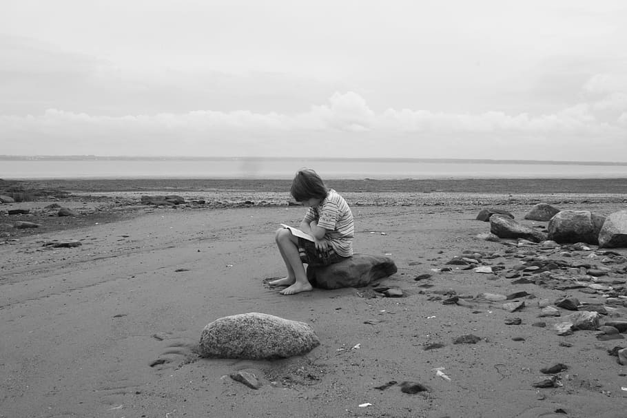 grayscale photo of girl reading book on beach while sitting on rock HD wallpaper