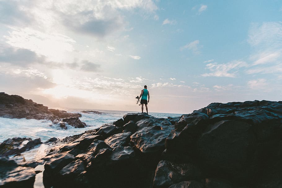 man standing on rocky shore under cloudy sky, person standing on big rock beside body of water under blue and white sky during daytime HD wallpaper