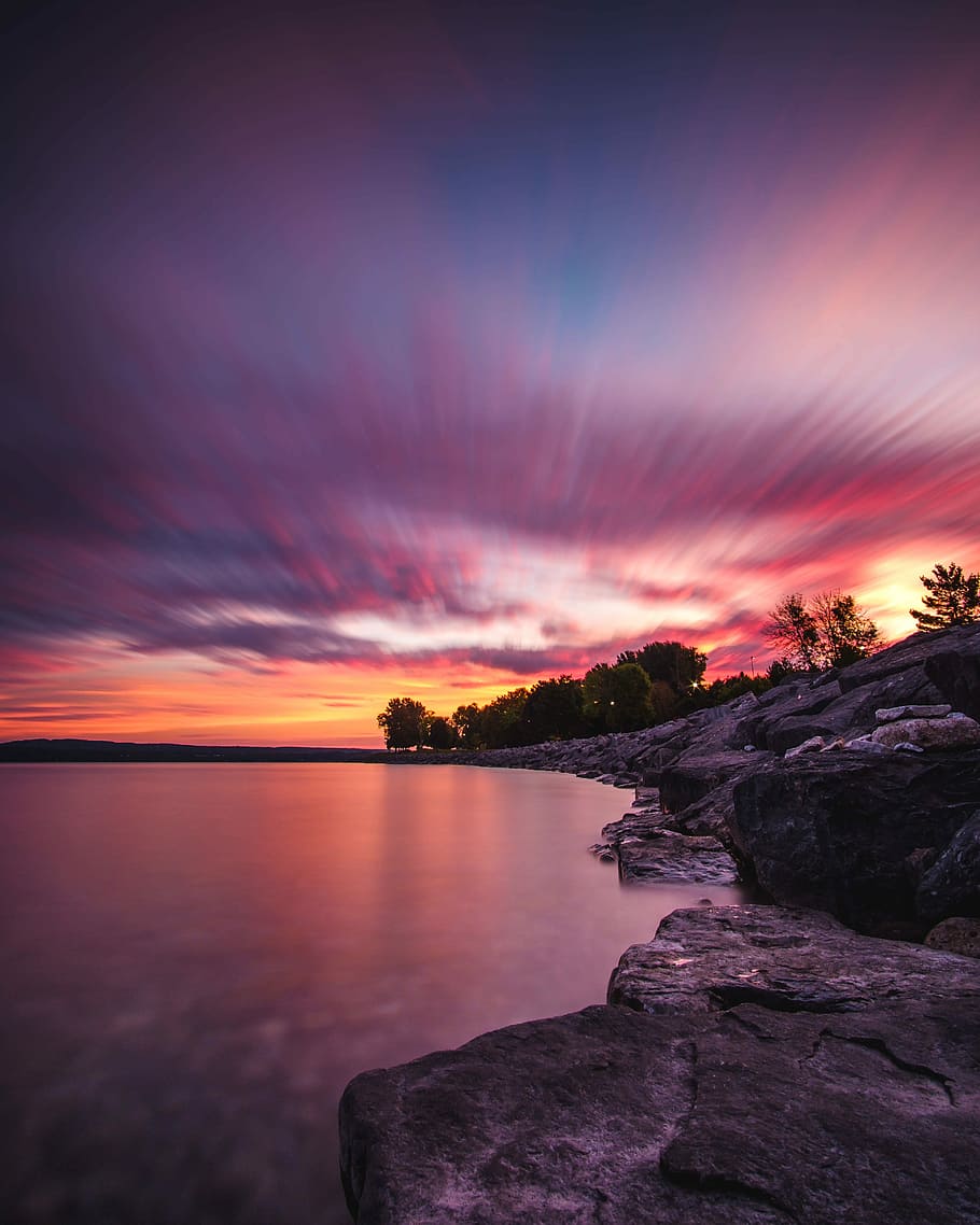 body of water and gray rock formation, timelapse photography of rocks beside body of water under red and gold skies HD wallpaper