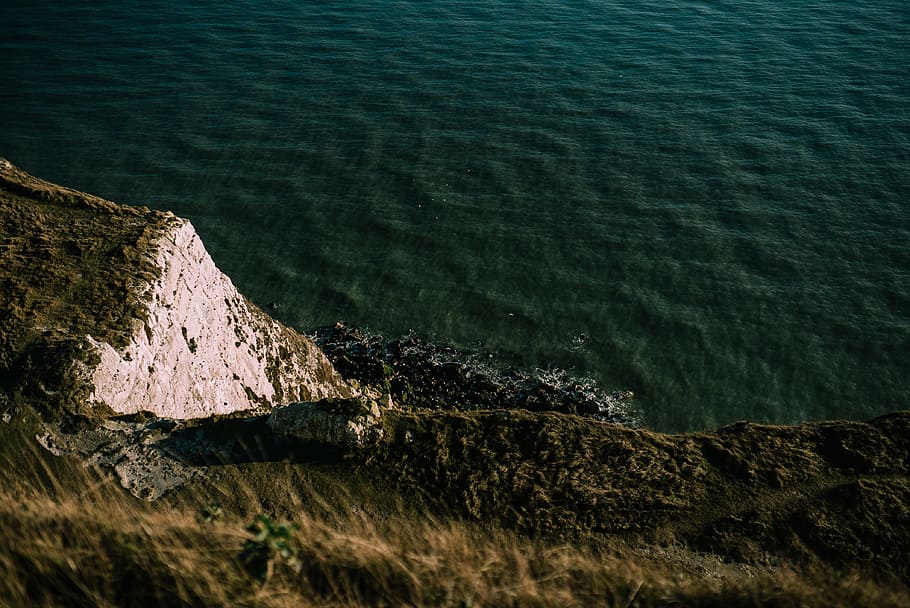 green grass cliff beside green body of water, rock mountain near the sea at daytime HD wallpaper