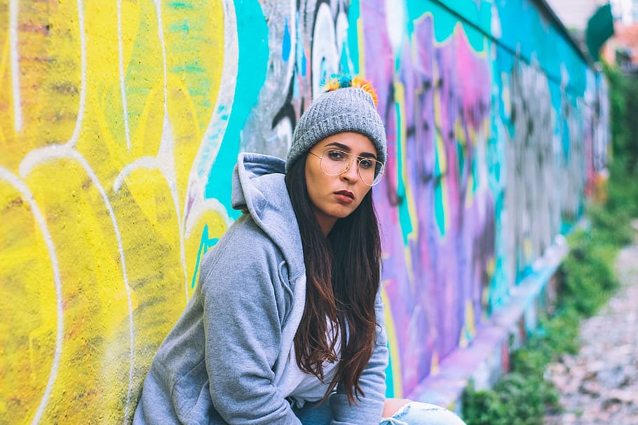 woman leaning beside wall during daytime, girl in gray jacket and bobble hat sitting on concrete wall with bench and painted graffiti at day time