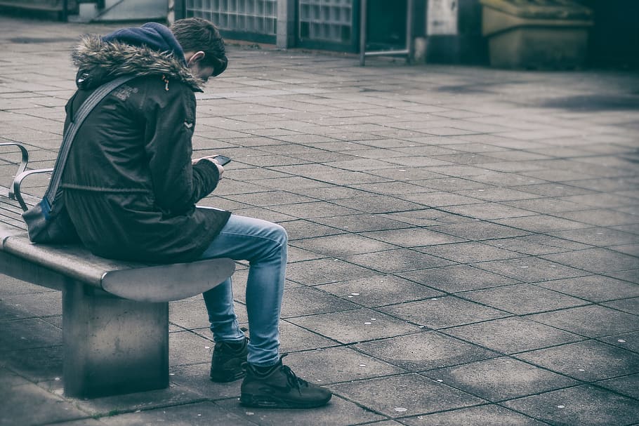 man sitting on bench holding phone, man in black jacket sitting on bench holding smartphone