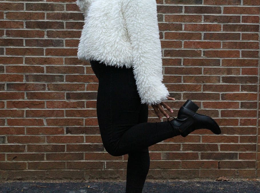 woman raising her left foot near brown brick wall during daytime, person wearing white fur jacket standing behind brown brick wall HD wallpaper