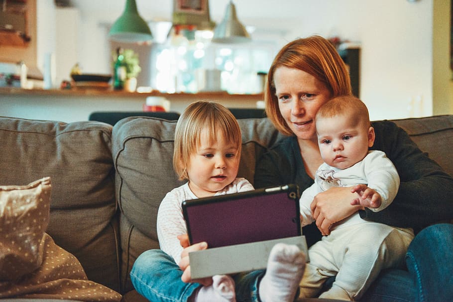 two babies and woman sitting on sofa while holding baby and watching on tablet, woman carrying baby beside girl holding table sitting on gray sofa