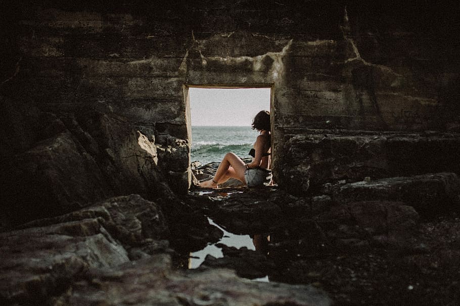 woman sitting inside cave, woman sitting on stone near body of water