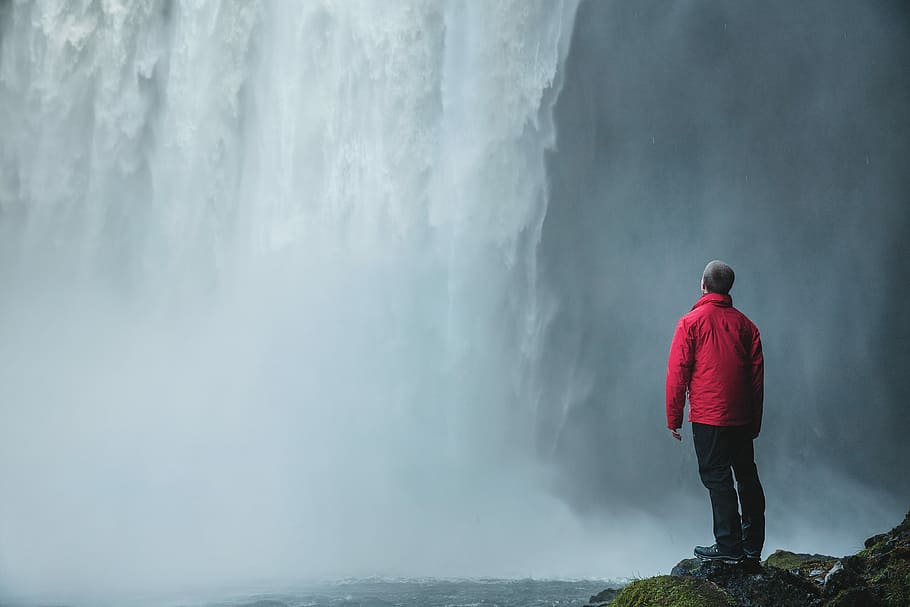man standing on a rock facing waterfalls, standing man watching waterfalls during day HD wallpaper