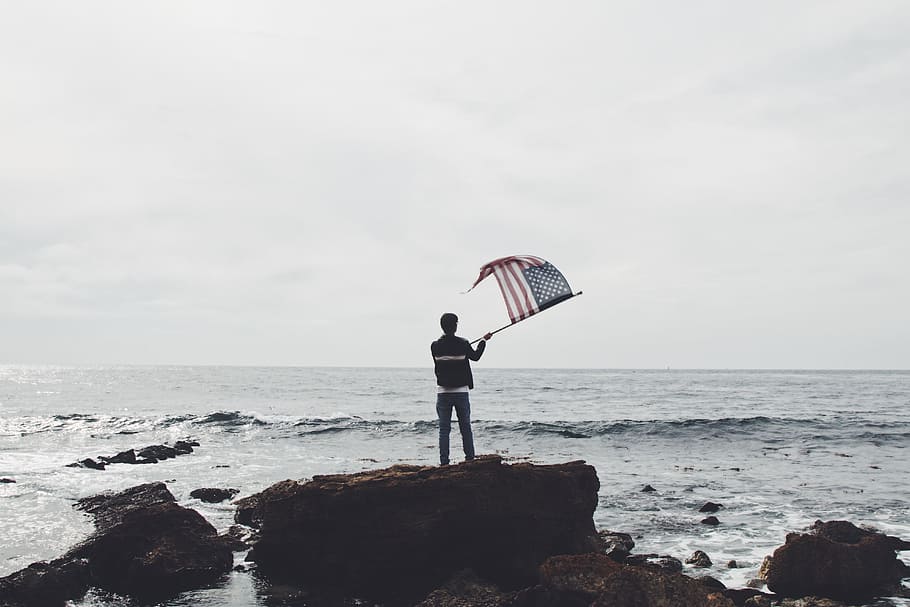 man holding flag of America standing on boulder near seashore, person holding American flag near on the body of water HD wallpaper