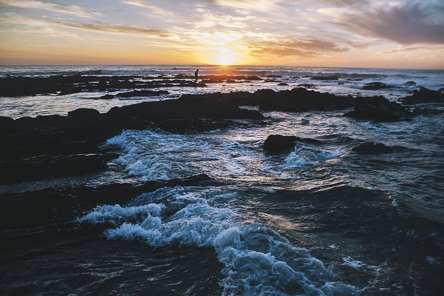 wave of water on rocks, person standing on rock seashore during golden hour HD wallpaper