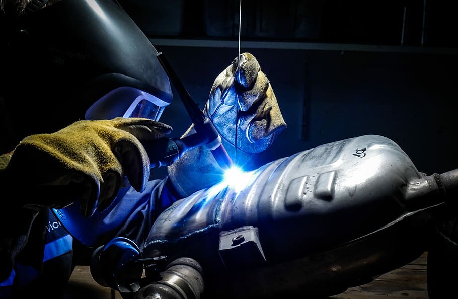 man wearing welding helmet welding on grey metal tank, person plasma welding gray vehicle gas tank