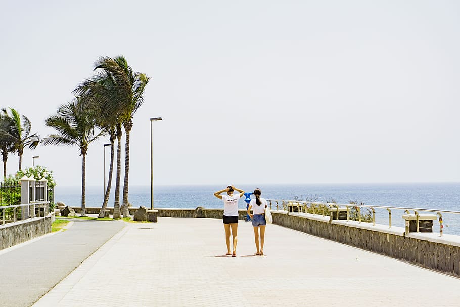 two women walking beside body of water, two women walking beside beach walk at daytime HD wallpaper