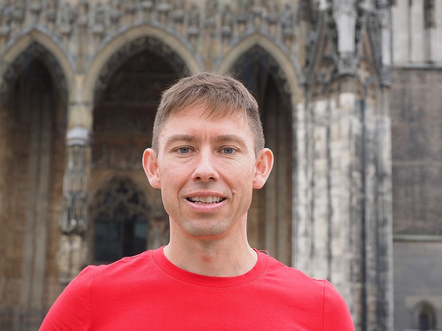man in red crew-neck shirt smiling beside grey concrete building HD wallpaper