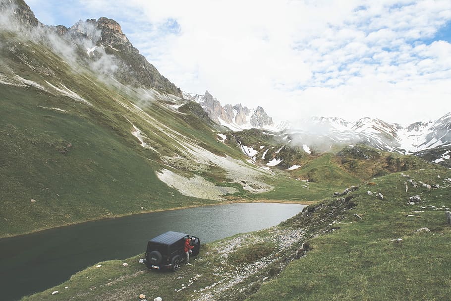 vehicle near body of water and person wearing red top under white and blue sky during daytime photography, black Jeep Wrangler parked near river and ice capped mountains during cloudy day