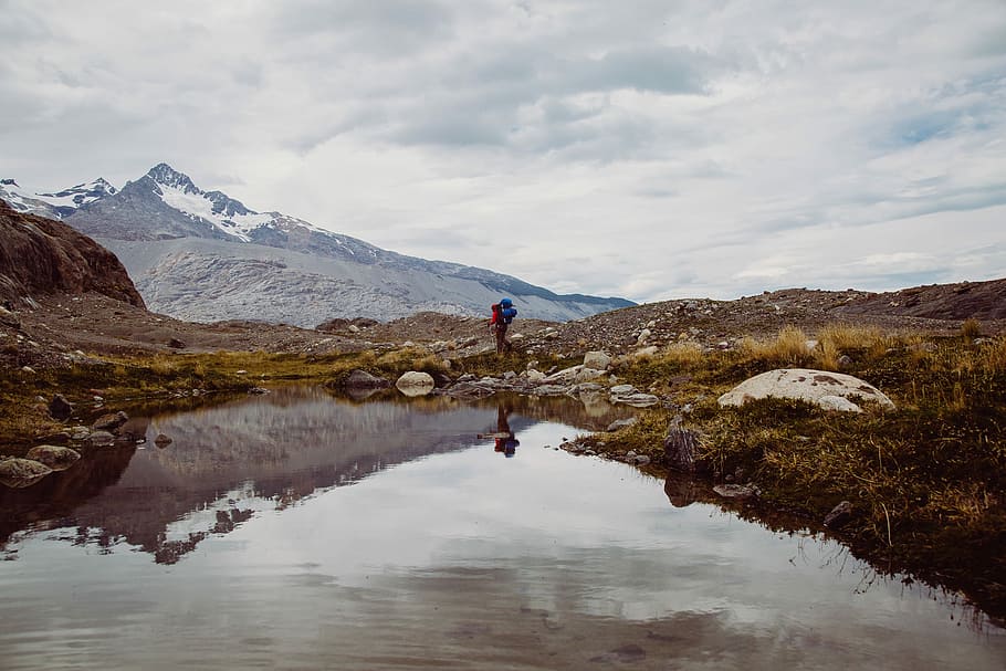 lake near mountain alps, man standing on brown sand with body of water during daytime HD wallpaper
