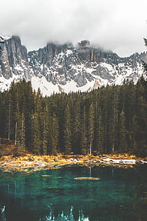 body of water near pine trees with snow-capped mountain at distance, rock formation covered with fogs near green trees and body of water at daytime HD wallpaper
