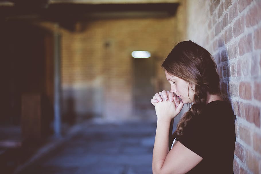 woman praying while leaning against brick wall, woman wearing black t-shirt