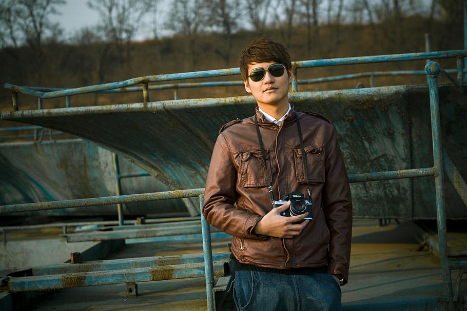 man wearing brown leather zip-up jacket holding black DSLR camera HD wallpaper