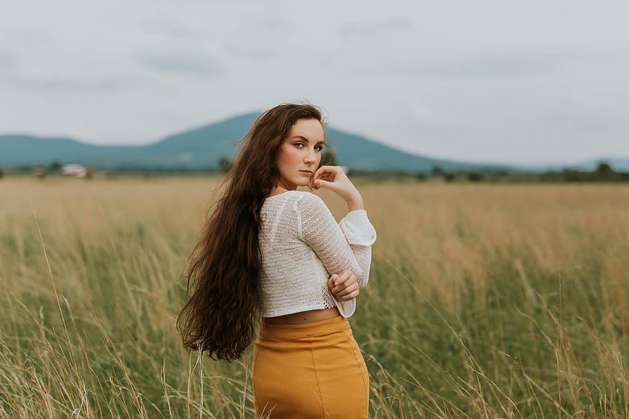 woman standing on grass field, woman wearing white sheer long-sleeved crop top and yellow bodycon skirt standing in middle of grass field