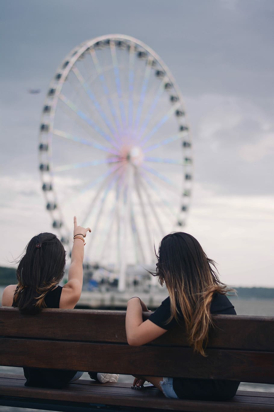 two women sitting on bench pointing white and blue Ferris wheel, two women in black top sitting on wooden bench fronting ferris wheel, HD wallpaper