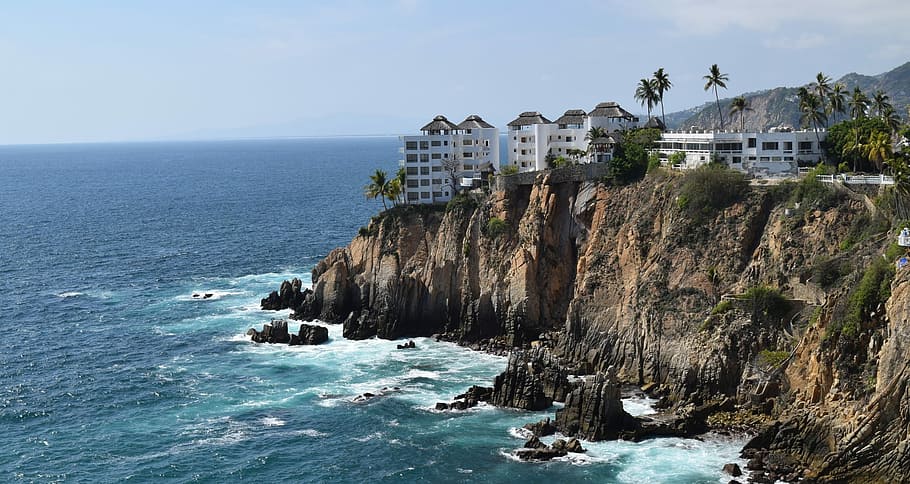 white concrete building surrounded by palm trees on cliff near sea at daytime HD wallpaper